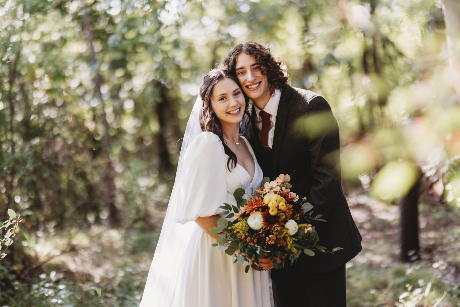 A bride and groom pose together in a sunlit forest setting with a vibrant autumn bouquet.