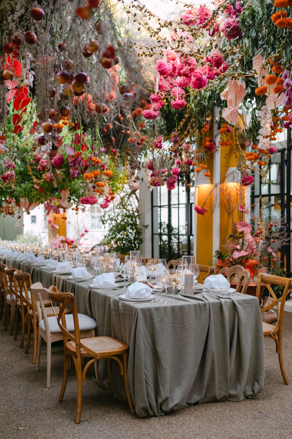 Elegant outdoor dining table with gray tablecloth under a canopy of hanging pink and orange flowers and greenery.