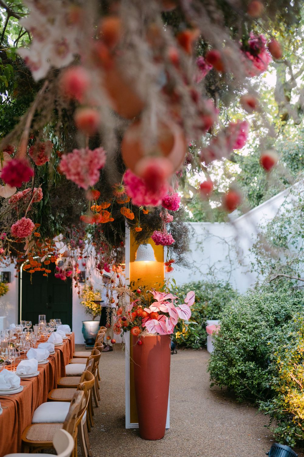 Elegant outdoor dining setup with hanging pink flowers and orange lanterns along a rustic pathway creates a romantic ambiance.
