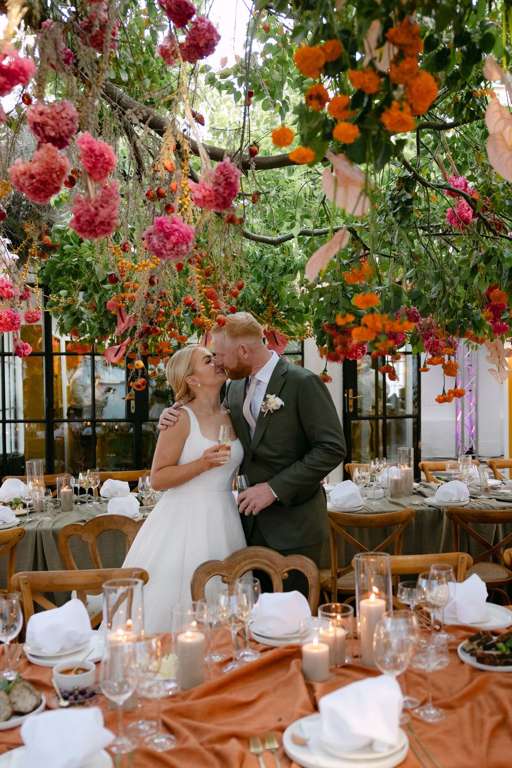 Wedding reception tables decorated with hanging pink and orange flowers and glowing candles create a romantic atmosphere.