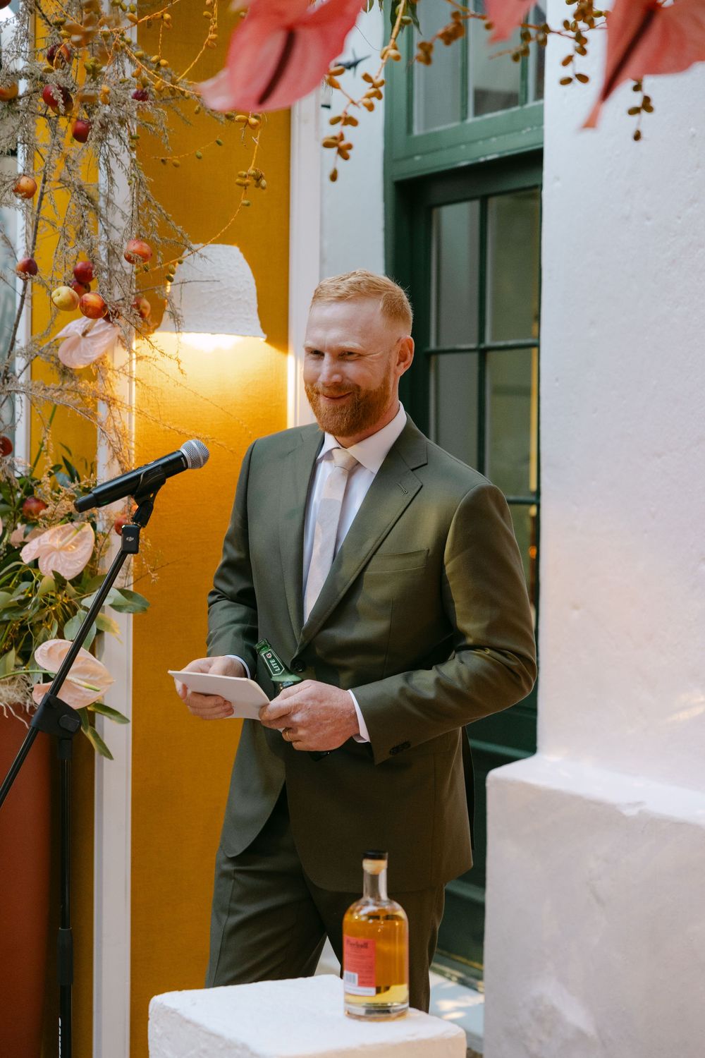 A speaker in a dark suit delivers remarks at a decorated venue with floral arrangements and warm lighting.
