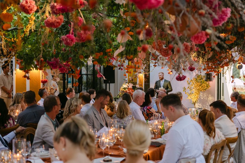 Wedding guests dine beneath vibrant floral decorations and glowing candles at an elegant outdoor reception.
