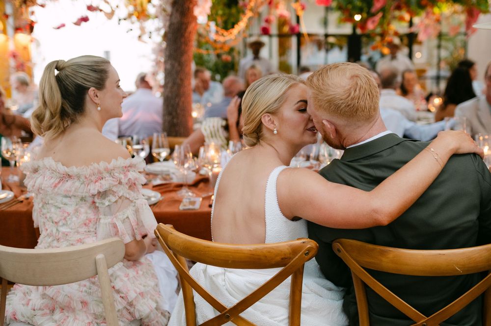 Wedding guests sit together in wooden chairs at an outdoor reception venue decorated with autumn colors and foliage.