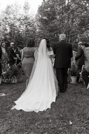A black and white photograph of an outdoor wedding ceremony with a bride in a flowing veil walking down the aisle.