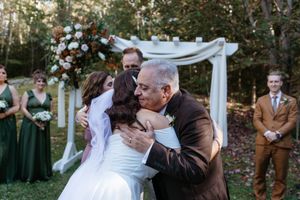 An outdoor wedding ceremony under trees with emerald green bridesmaids dresses and white floral arch.