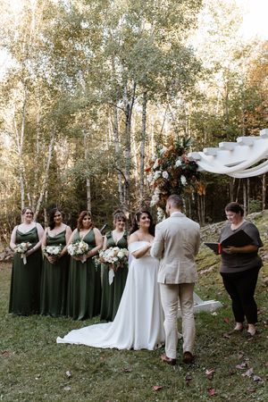 Wedding party in black dresses and tan suits poses together on grassy hill with autumn trees in background.