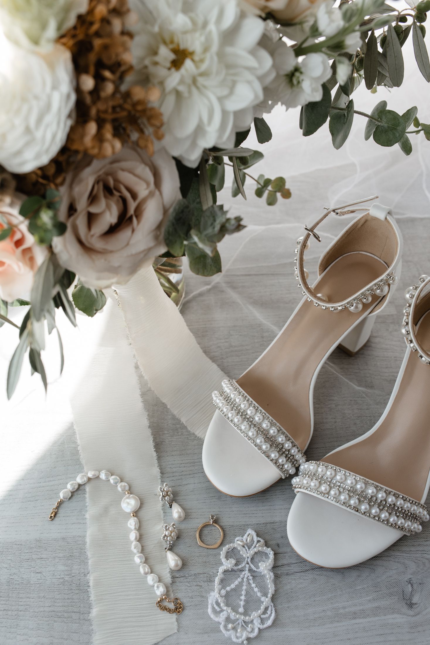 Wedding accessories including white heels, pearl jewelry, rings and delicate floral arrangements on a light gray surface.