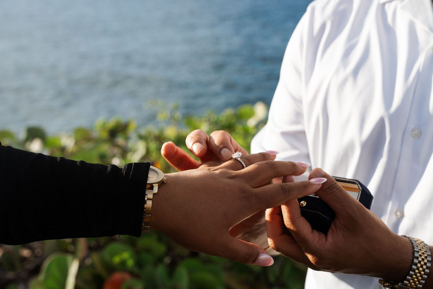 Hands of a couple in white clothing hold each other tenderly against a blurred outdoor background.