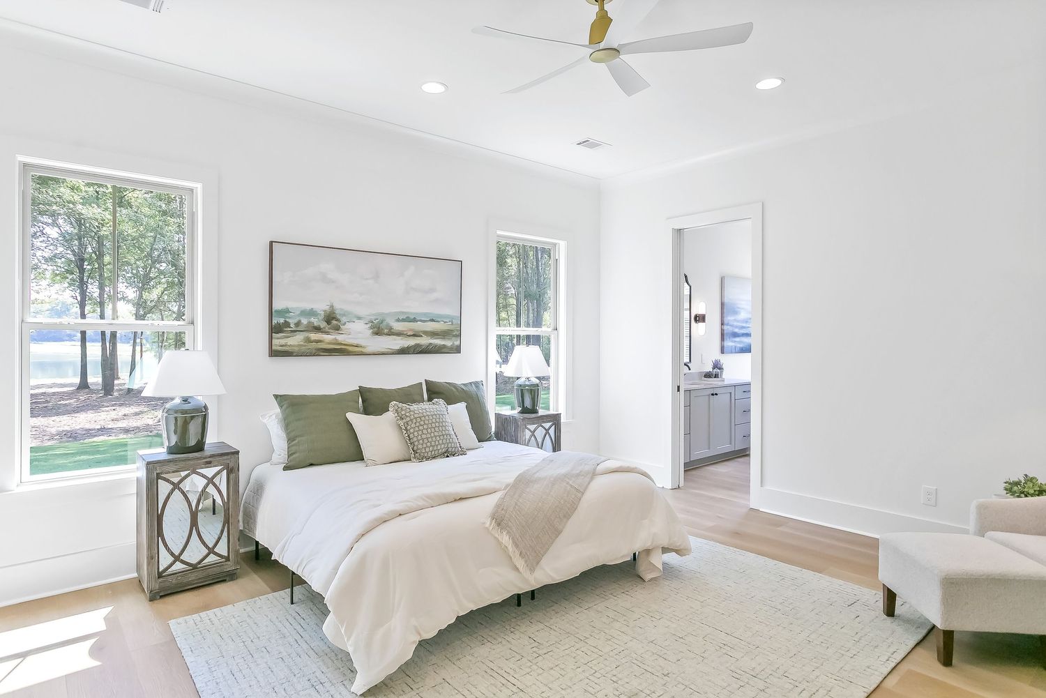 Modern white bedroom interior with ceiling fan, green accent pillows, and large windows letting in natural light.
