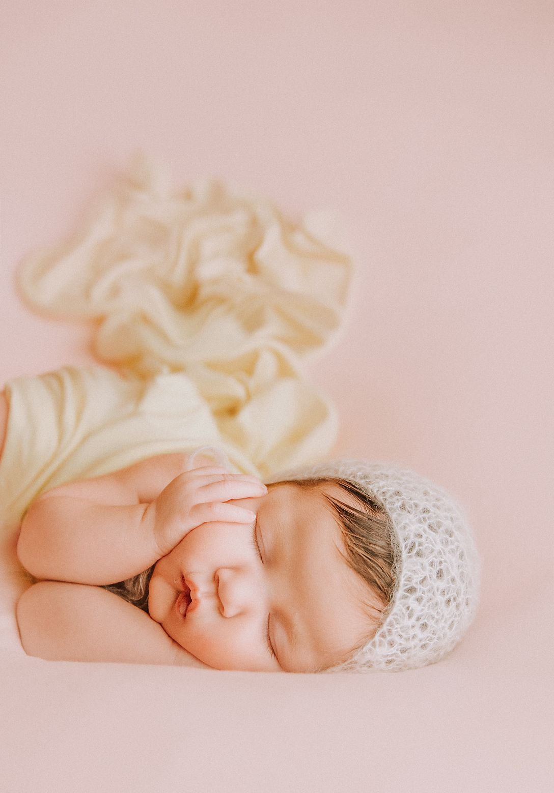 A sleeping newborn baby wearing a delicate lace bonnet rests peacefully on soft cream-colored fabric in natural light.