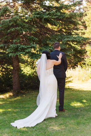 A wedding couple shares a romantic moment in a sunlit garden with flowing white veil and evergreen trees in the background.