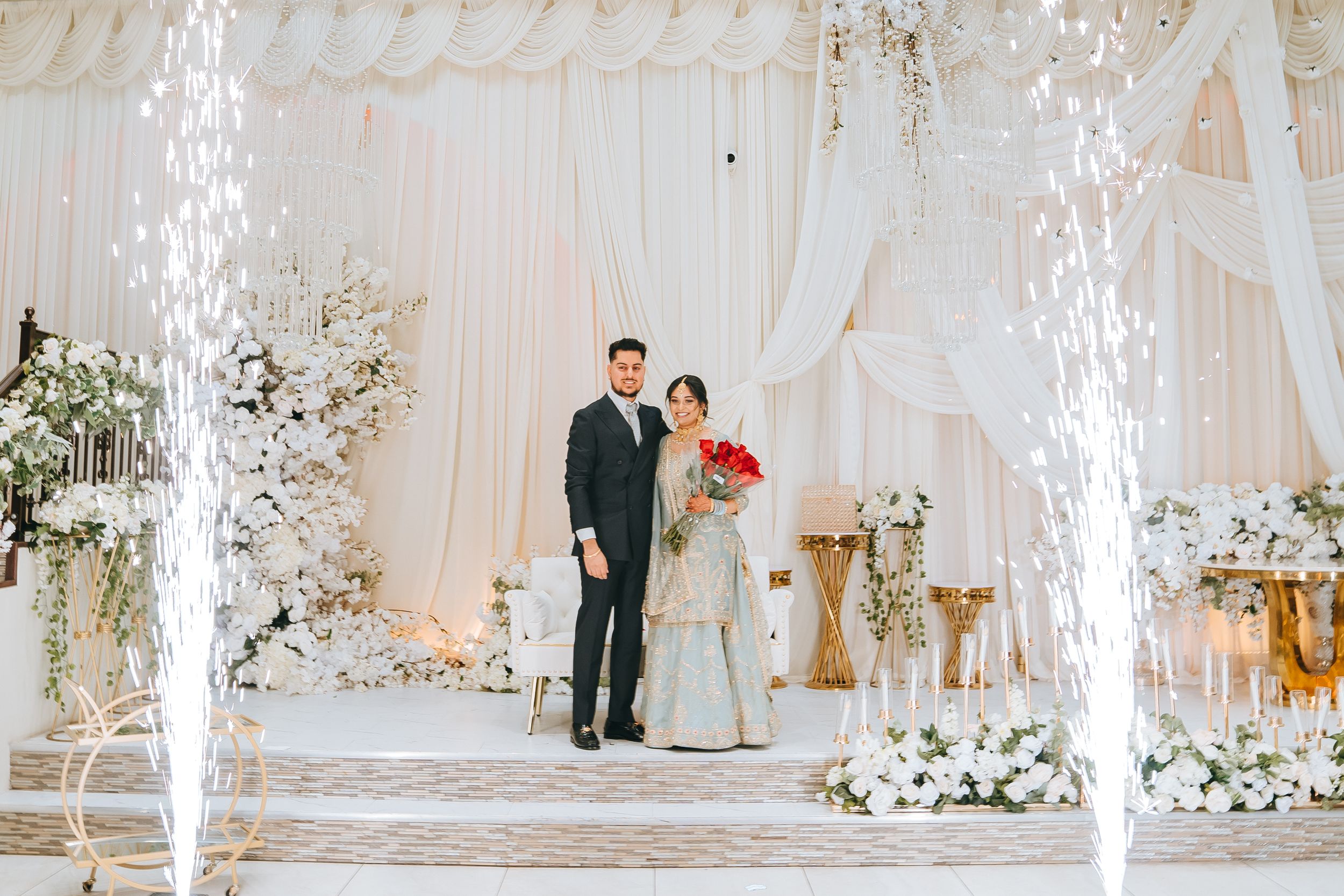 An elegant wedding reception setup with white drapes, chandeliers, and floral arrangements.