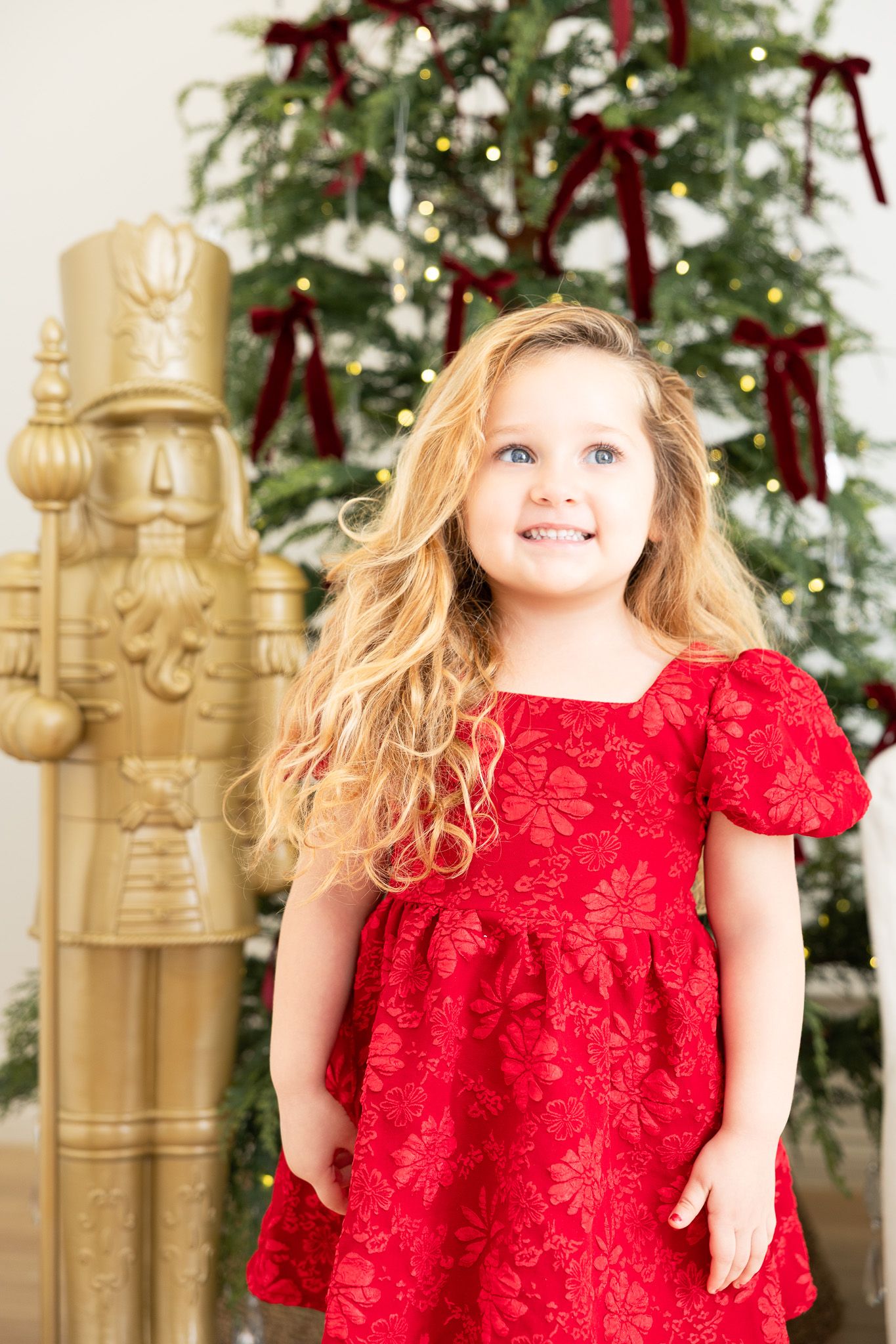 A young child in a red velvet holiday dress twirls and plays in front of a decorated Christmas tree and gold decor.