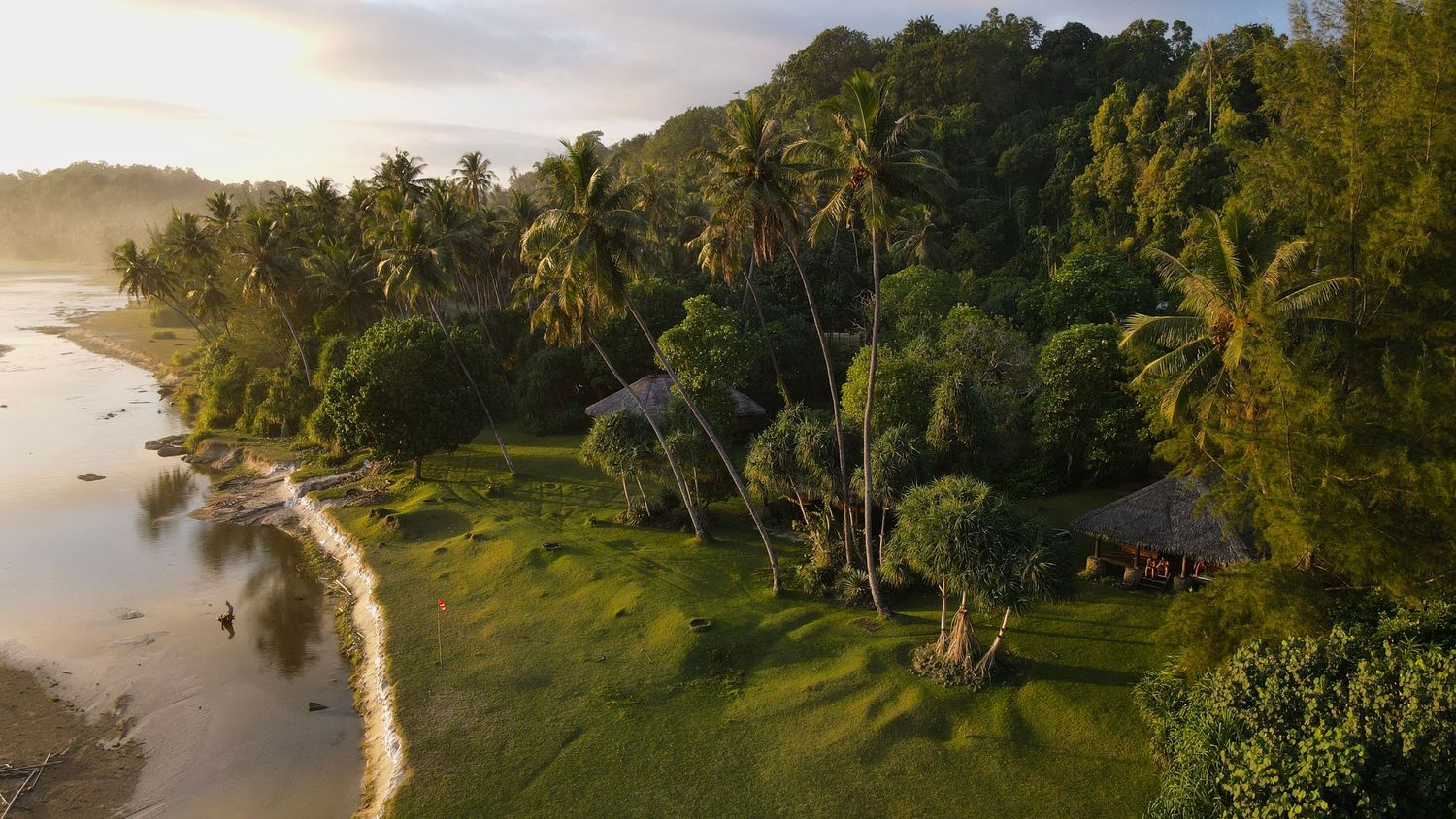 Aerial view of a tropical coastline with palm trees and lush greenery along a curved beach at sunset.