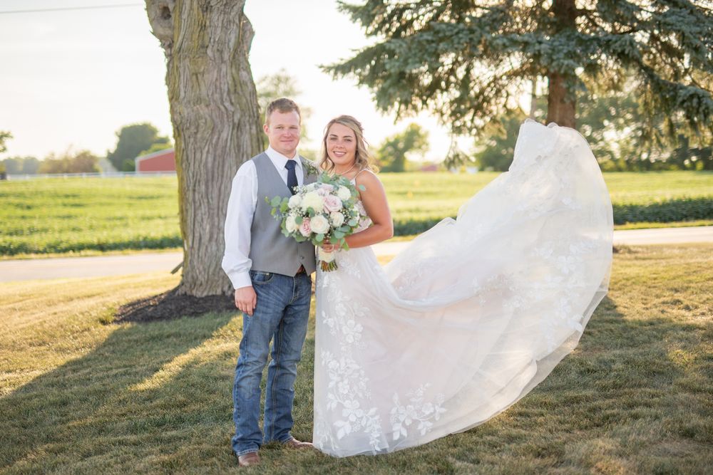 Wedding couple poses under large trees in a scenic outdoor garden setting.