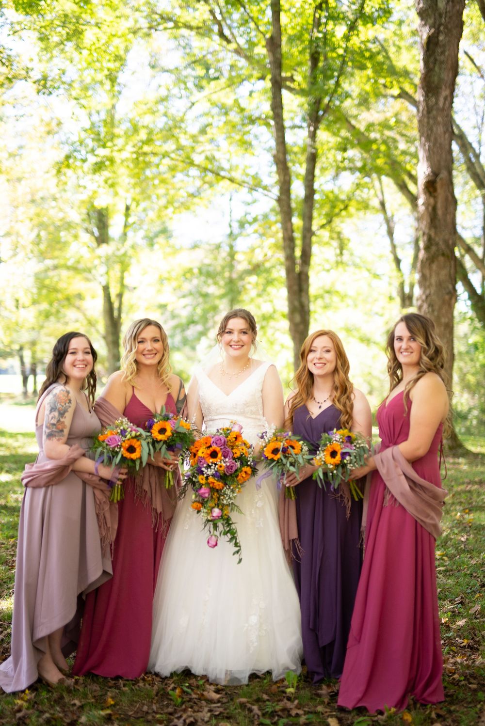 Bridesmaids in shades of burgundy and mauve dresses hold colorful autumn bouquets in an outdoor woodland setting.