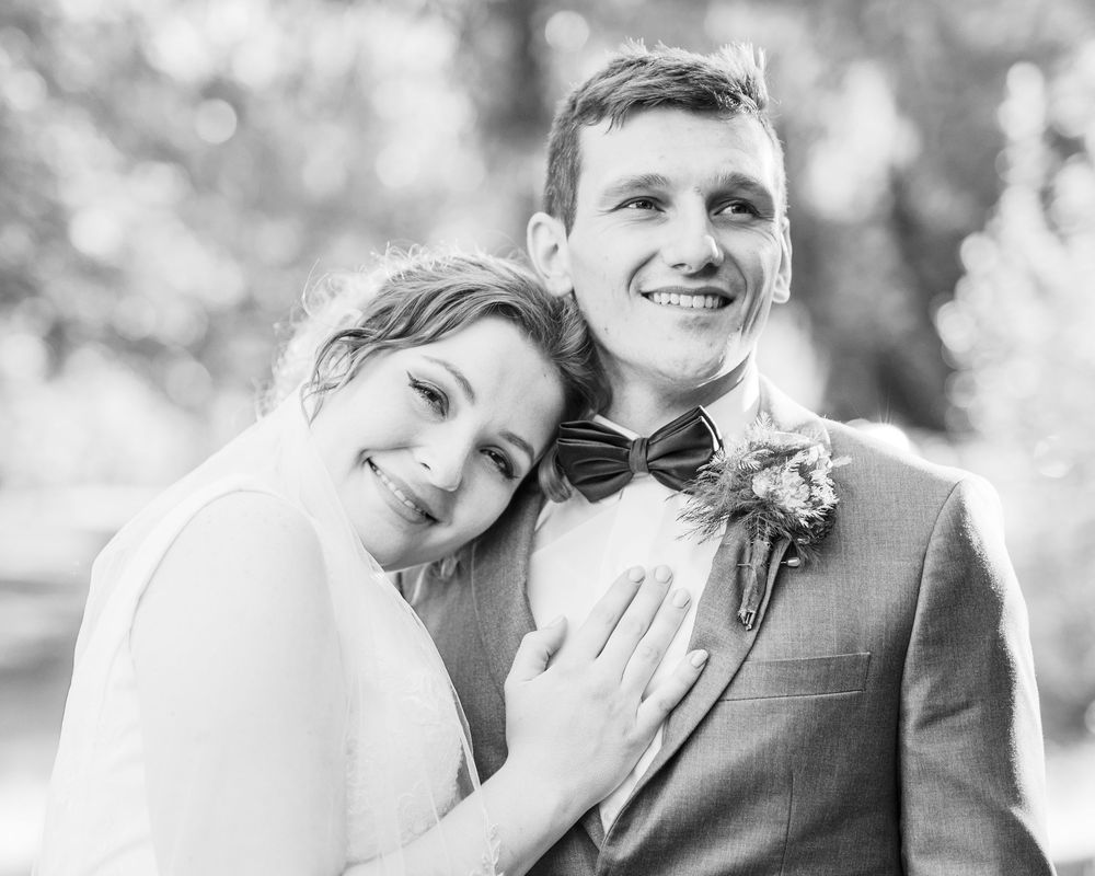 A joyful black and white portrait of a newlywed couple embracing outdoors, with the groom wearing a bowtie and boutonniere.