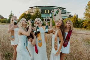 A group of graduates in white dresses celebrate with champagne in front of UO stadium.