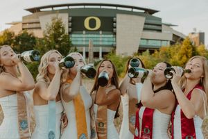 Graduates in white dresses and colorful stoles drink champagne during a celebratory photoshoot.