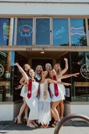 Group in matching white dresses and red ties pose playfully outside bar with vintage beer signs.