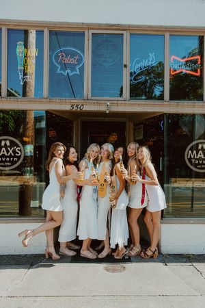 Group in coordinated white outfits pose with drinks outside bar featuring neon beer signs.