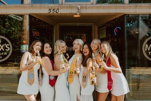 Friends in matching white dresses and red sashes pose with drinks outside vintage storefront.