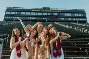 Group wearing white dresses and red sashes pose cheerfully in front of stadium bleachers with Welcome sign.