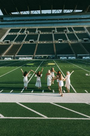 Group in white dresses celebrating with raised arms on football field with empty stadium seats behind.