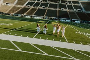 People in white dresses celebrate on football field with stadium seating in background.