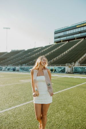 A person in a white dress poses playfully on a football field with stadium seating in the background.