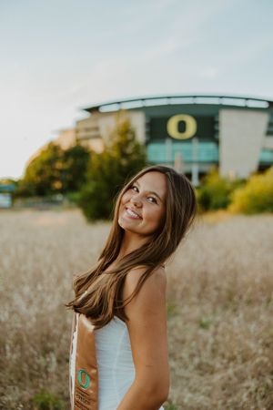 A graduate in white dress poses joyfully in a field with Oregon stadium in the background.