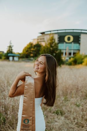 Graduate posing with gold stole in front of Oregon Ducks stadium during golden hour.