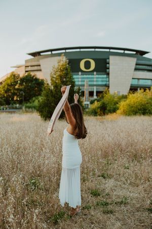 Graduate in white dress holding stole aloft at University of Oregon stadium.