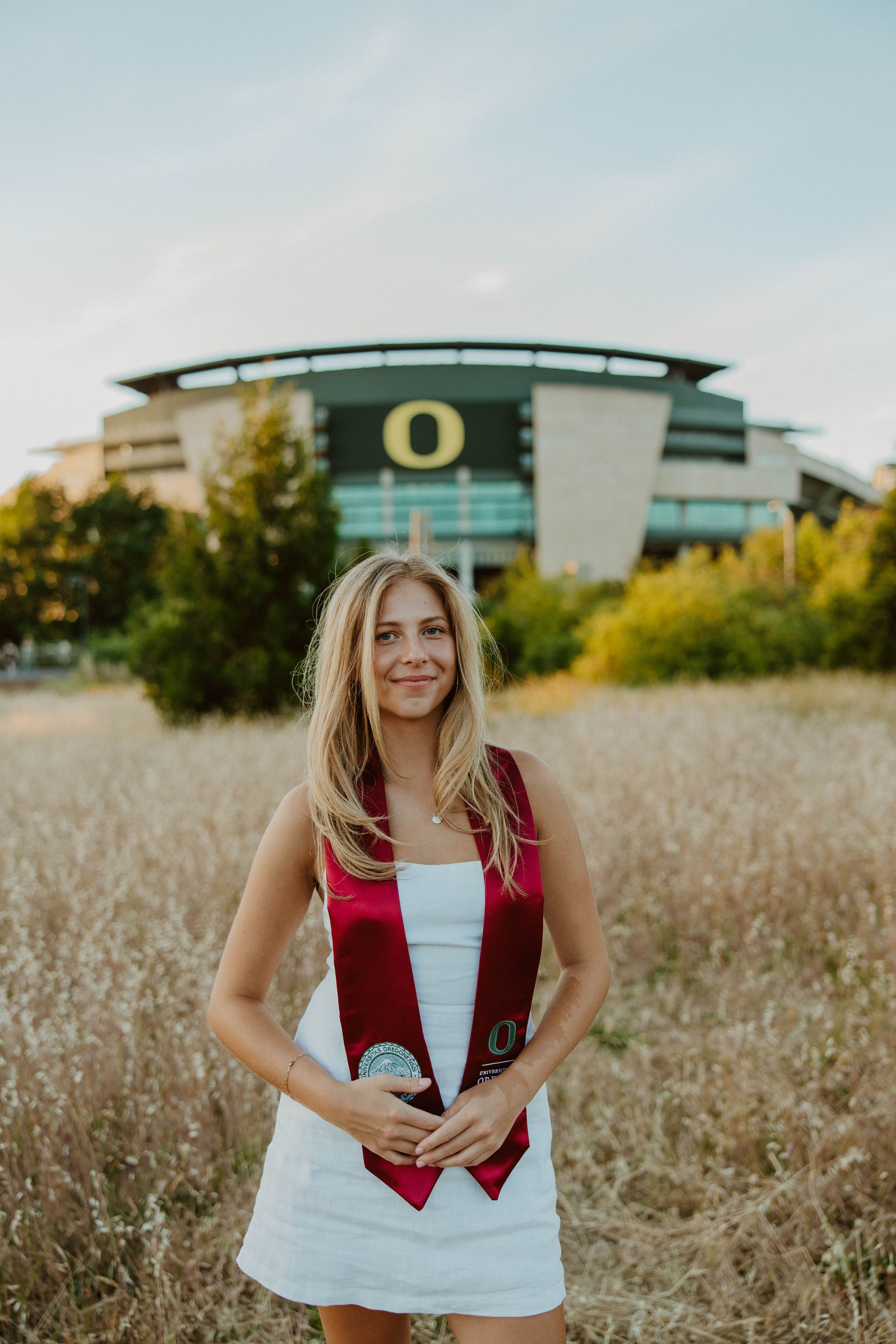 Graduate wearing red academic stole poses in field near Oregon Ducks stadium.