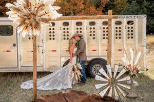 Newlyweds pose by rustic horse trailer decorated with dried pampas grass and boho wedding decor.
