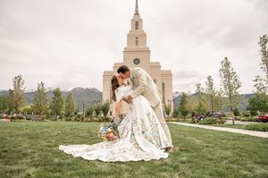 Wedding couple embracing on grass lawn with white church and mountains in background.