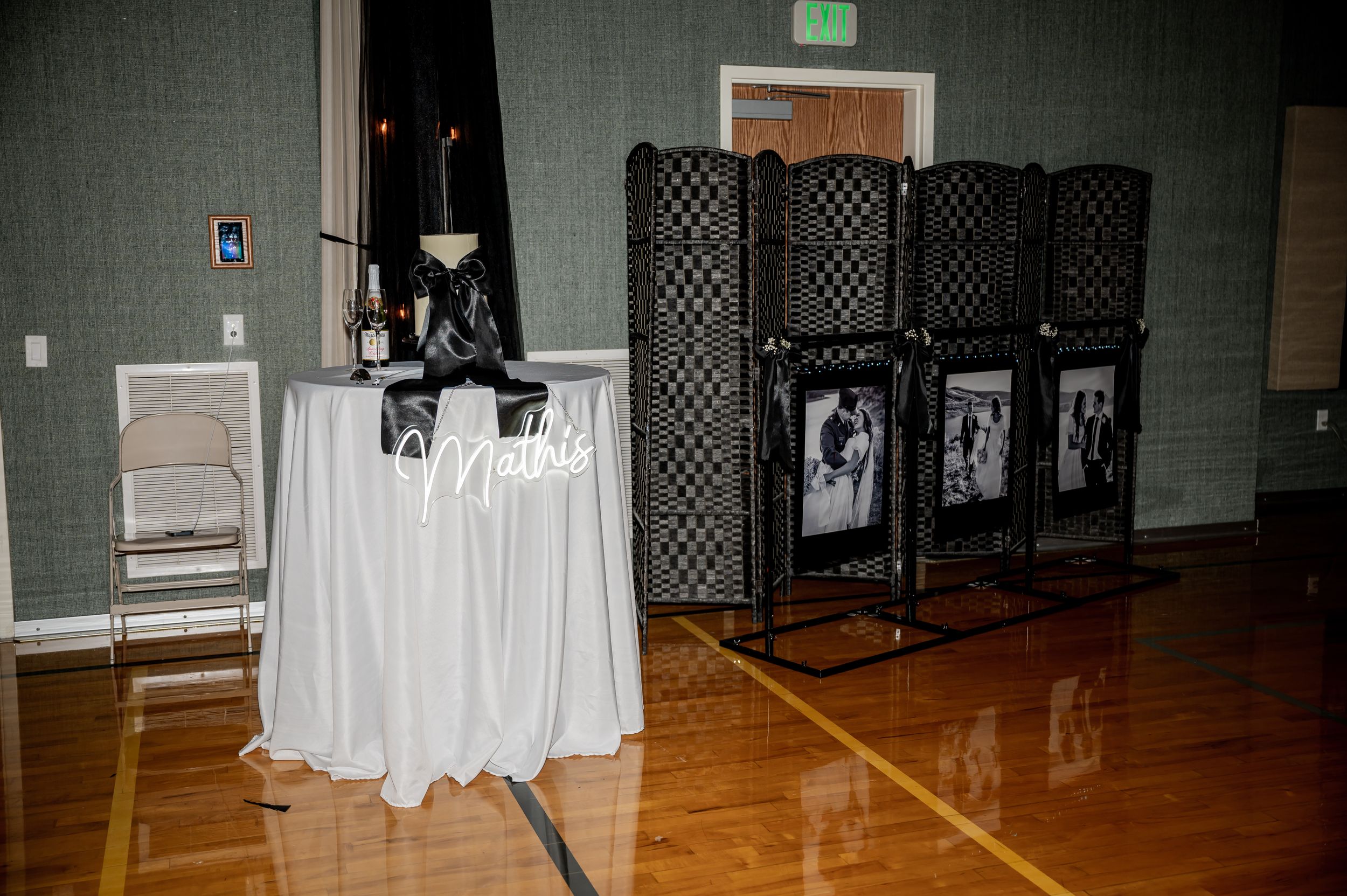 Wedding reception setup with white tablecloth and black room dividers displaying photos in a gymnasium.