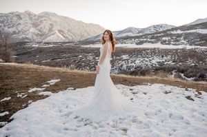 Bride in white gown poses on snowy mountain landscape at sunset.