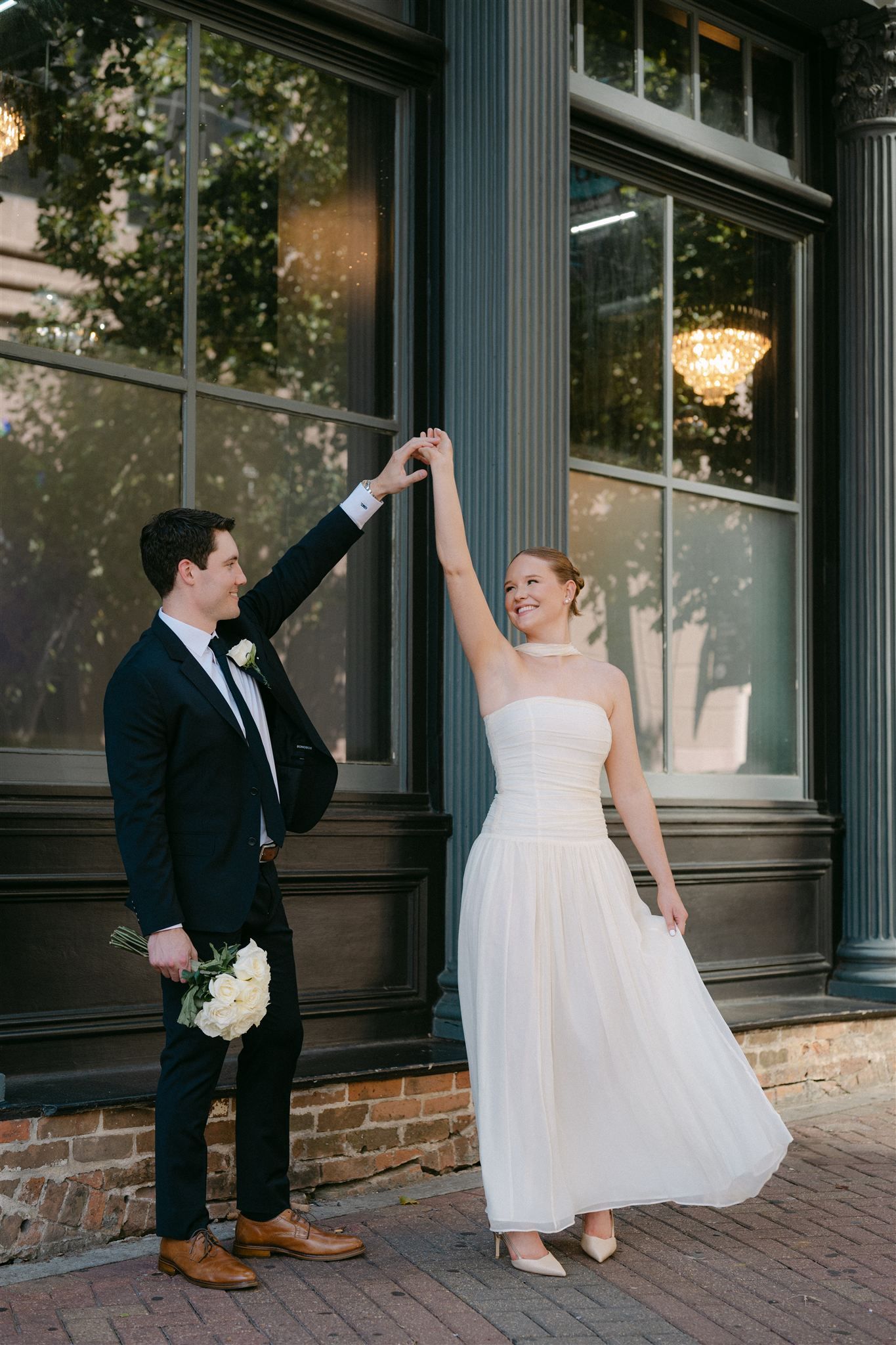 A couple in wedding attire share a romantic moment outside a historic building with elegant windows and chandeliers.