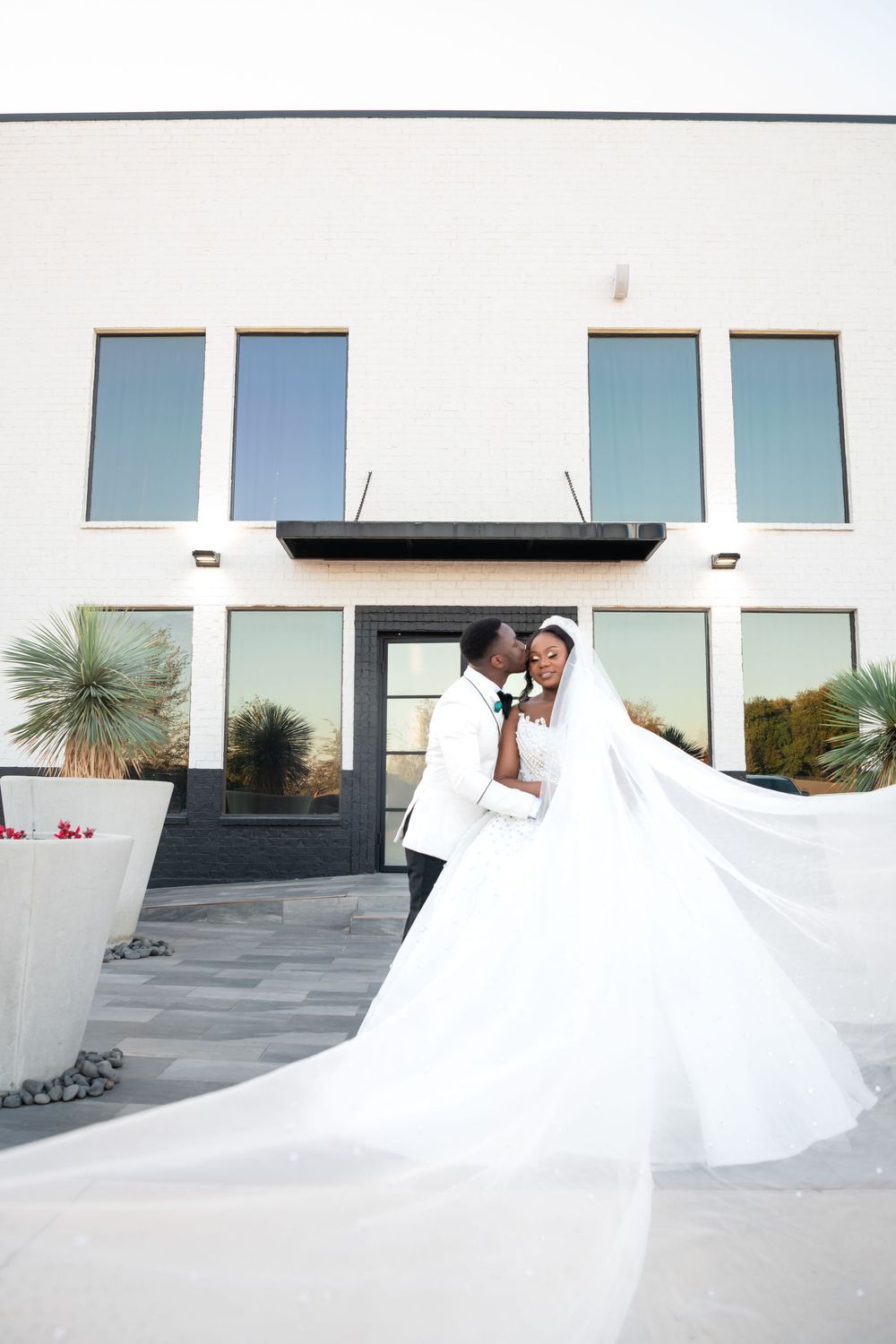 A flowing white wedding dress billows dramatically in the wind against a modern architectural backdrop with large windows.