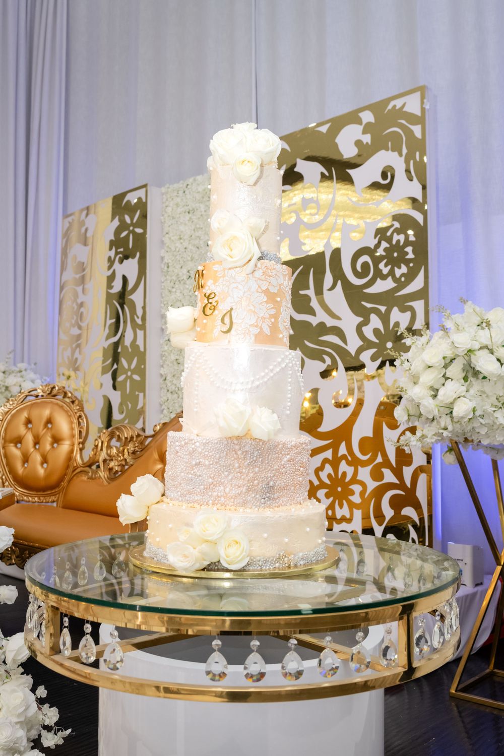 Elegant five-tier white and gold wedding cake displayed on glass stand with decorative backdrop.