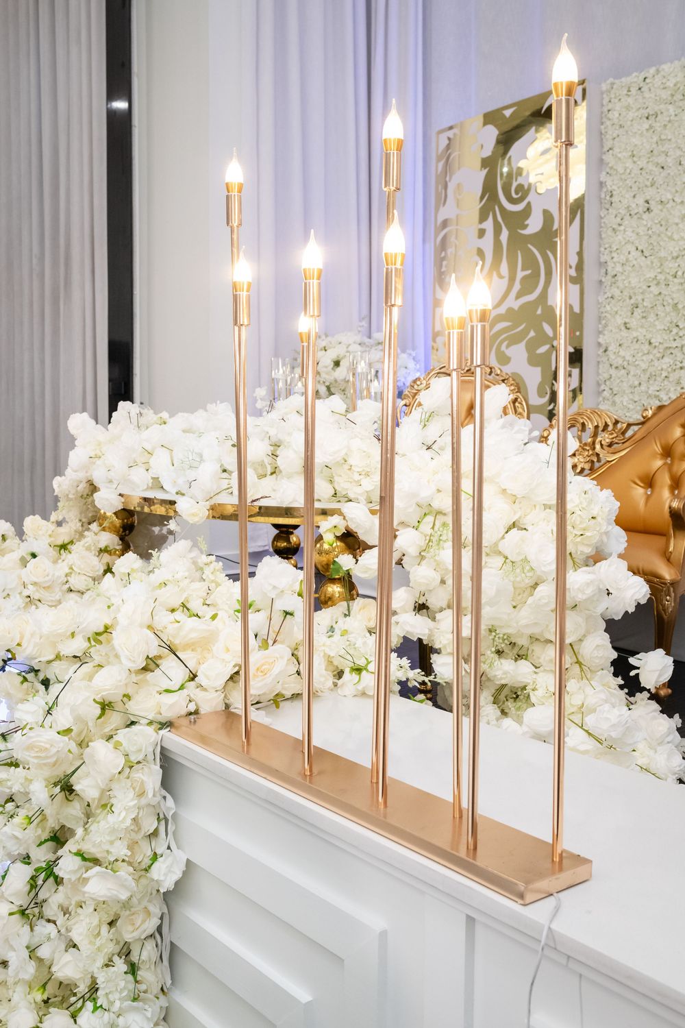 Elegant gold candlesticks with white floral arrangements line a mirrored wall in a luxurious event setting.