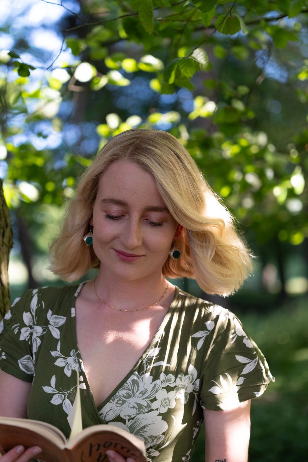 A woman in a floral dress sits outdoors under dappled sunlight and green foliage.