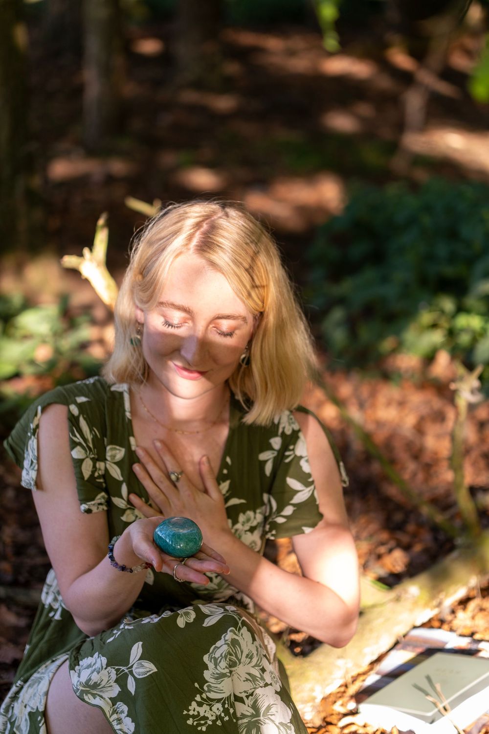 Person in floral green dress sits in dappled sunlight holding a blue crystal while relaxing outdoors.
