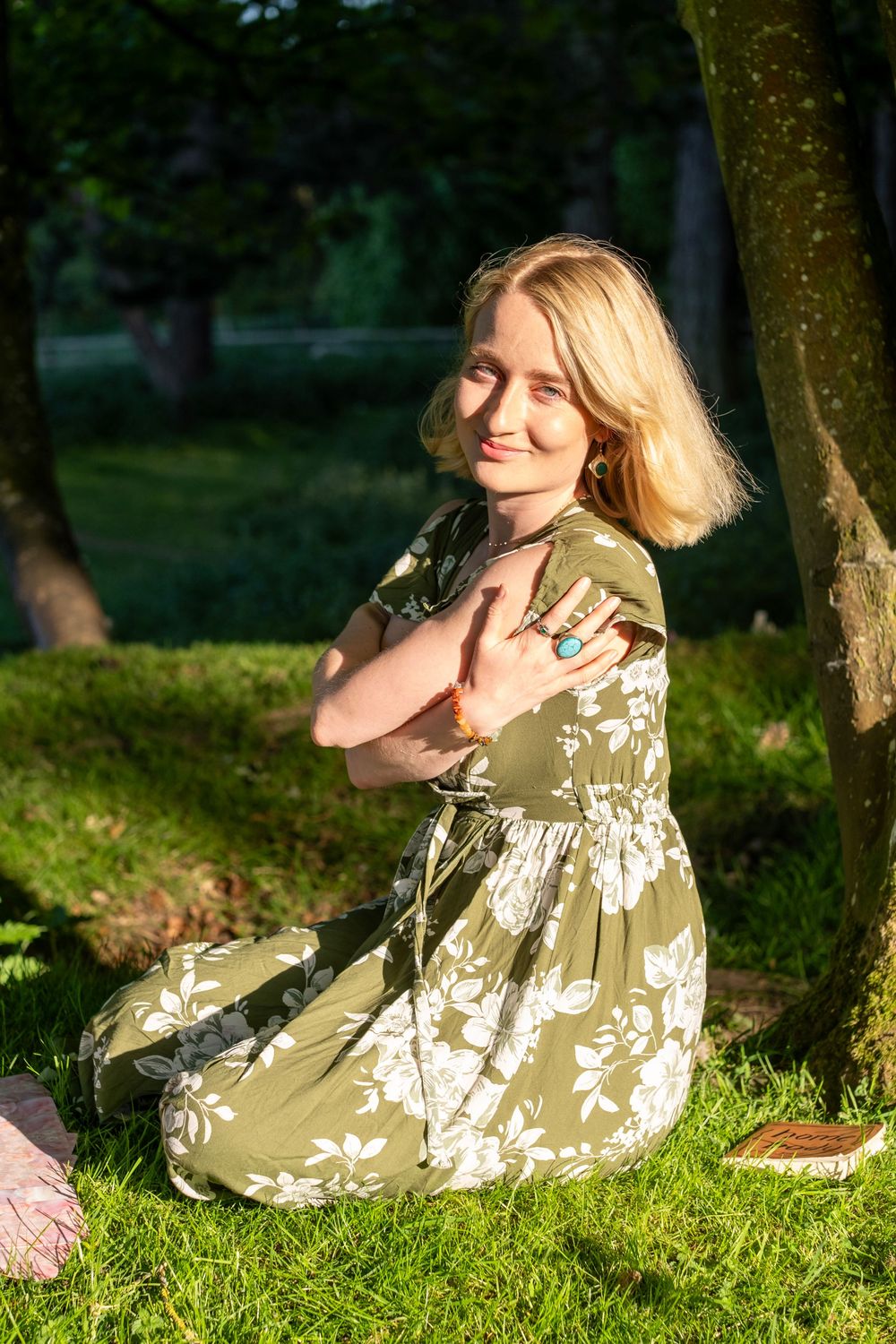 Woman in floral dress relaxing on grass in dappled sunlight beneath trees on a summer day.