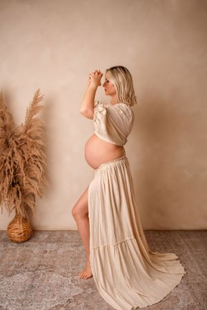 Maternity photo series in warm tones showing a flowing cream dress and pampas grass decoration against neutral backdrop.