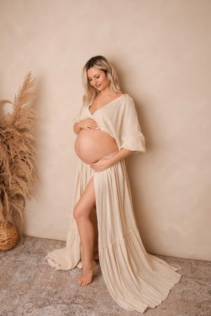 A maternity photoshoot featuring a flowing cream dress and pampas grass decor against a neutral beige backdrop.