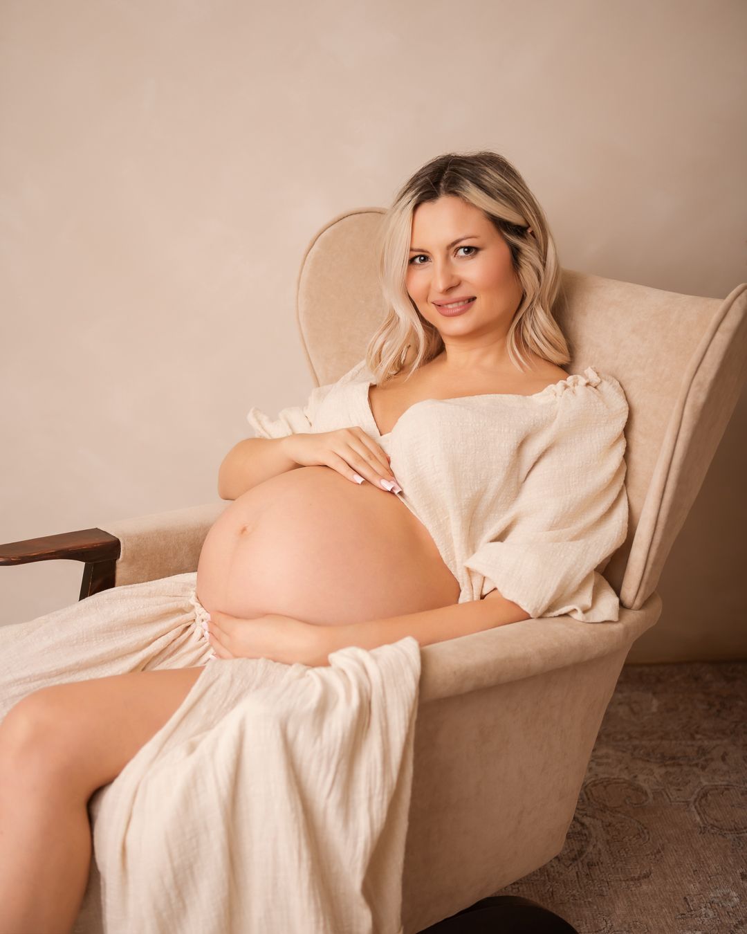 A maternity photoshoot series showing a pregnant woman in a white dress reclining on a beige couch against a neutral background.