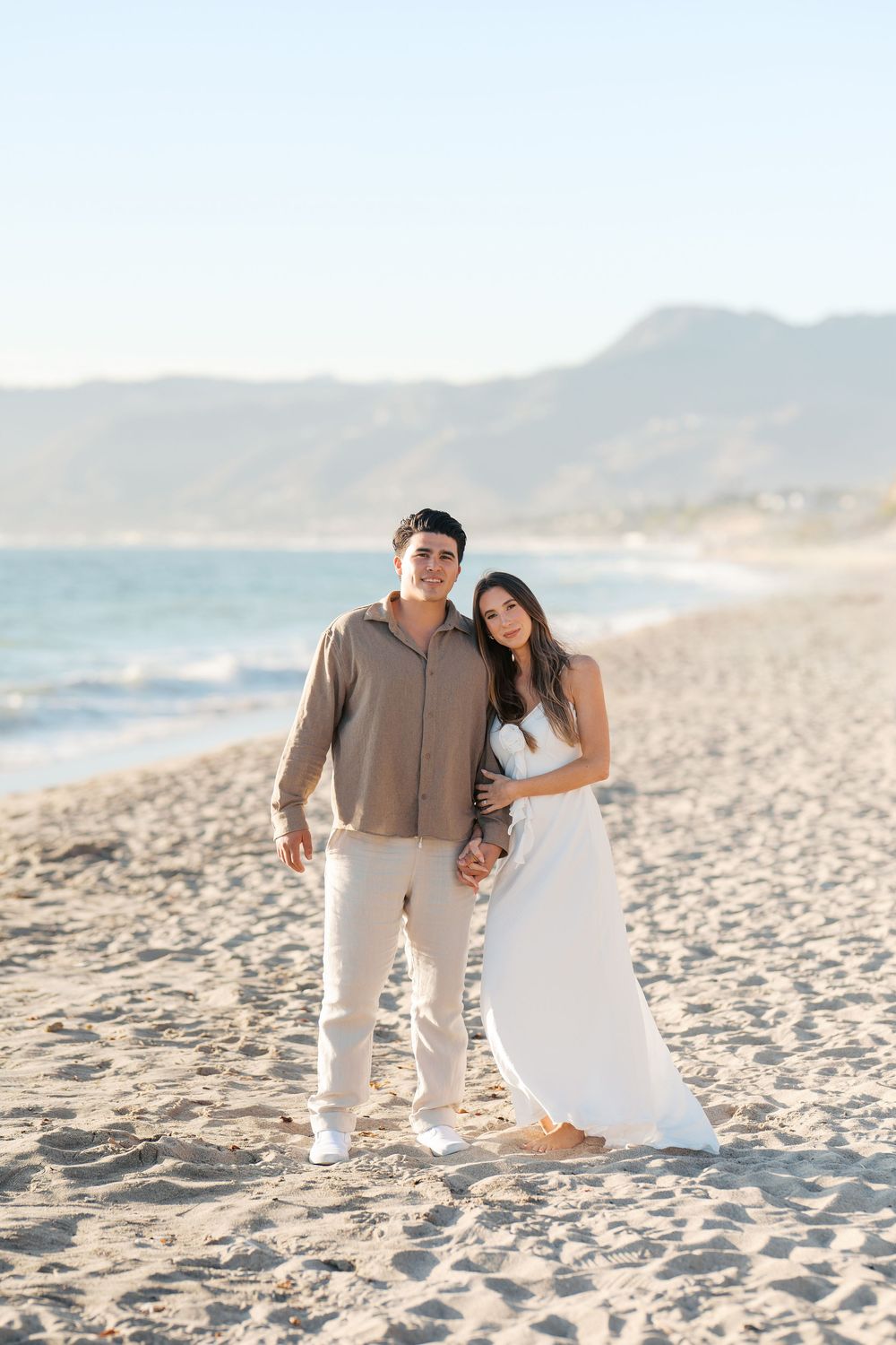 A couple walks together on a scenic beach at sunset, with mountains in the background and footprints in the sand.