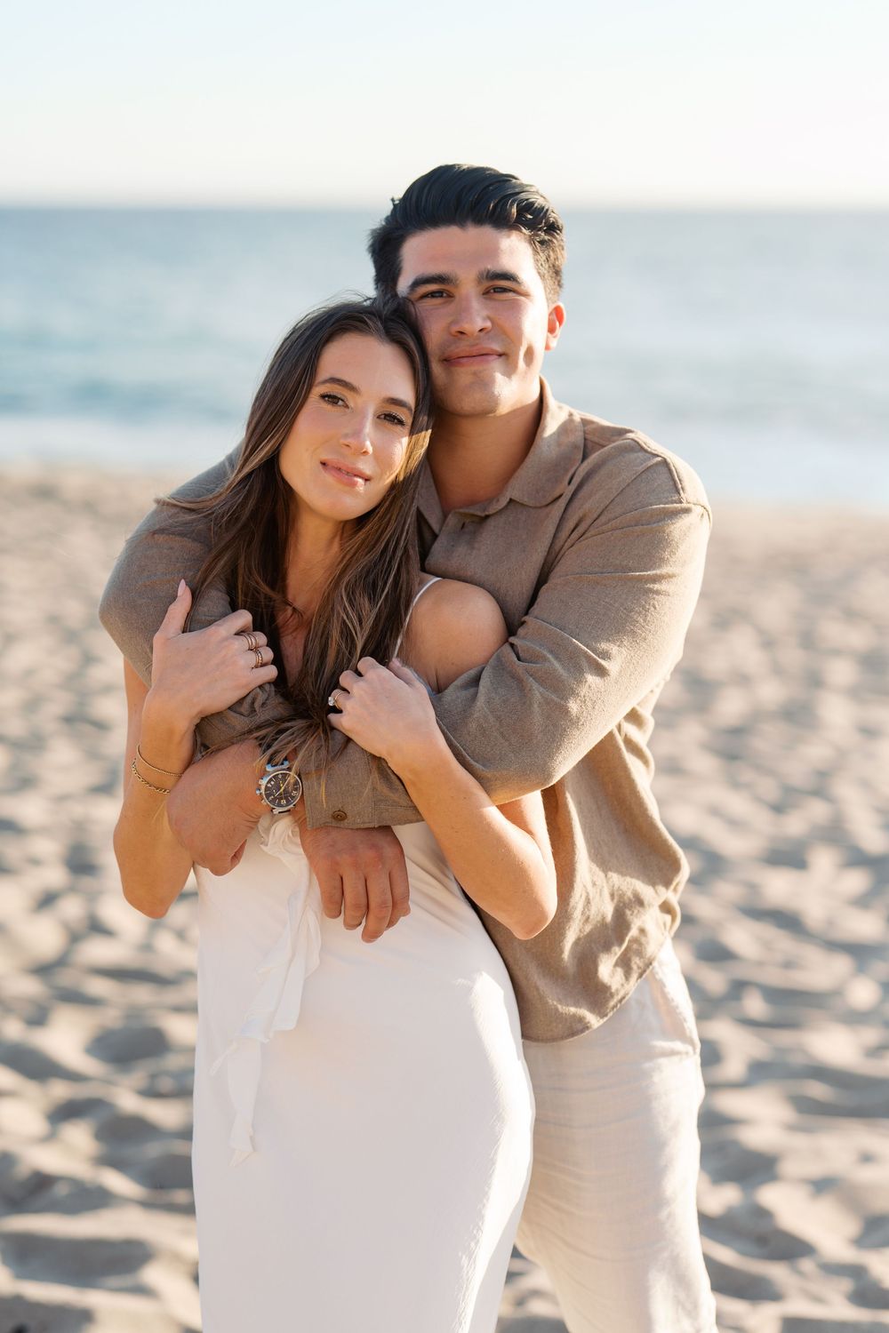 A romantic beach photoshoot featuring a couple embracing in casual attire against a sandy backdrop.